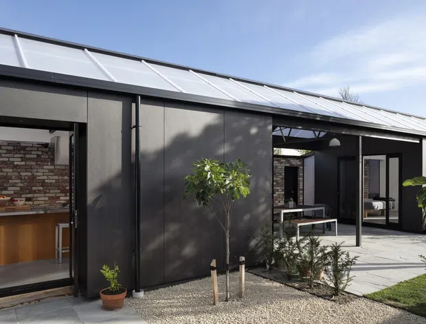 Contemporary single-storey home exterior arranged around a sheltered courtyard. The dark rendered or panelled facade has broad floor-to-ceiling openings, a light metal pitched roof with extended eaves, and a covered outdoor dining or lounge area. A partially visible kitchen with brick feature wall and timber cabinetry opens directly to the courtyard. Gravel landscaping, restrained planting, concrete paving, and black structural framing create a crisp modern aesthetic.
