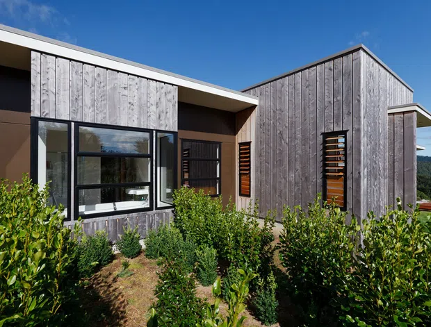 Exterior view of a contemporary single-storey building with a low-pitched roof and articulated gabled volume. The facade combines weathered vertical timber cladding, dark painted wall surfaces, black-framed glazing, and timber privacy screens. Dense low planting occupies the foreground, with wooded hills visible beyond.