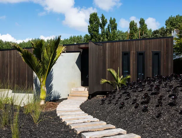 Single-storey contemporary home exterior with dark vertical timber cladding, a flat roof, and a contrasting smooth light-grey rendered entry volume. A curving stepping-stone path leads through a dark landscaped garden with palm-like and ornamental plantings. Tall narrow windows and strong horizontal volumes create a minimalist modern facade under a bright blue sky.