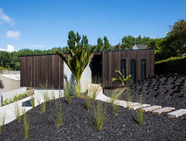 Exterior view of a low-slung modern residence composed of dark vertical timber-clad volumes set into a landscaped sloping site. Pale concrete paths and retaining walls contrast with black gravel, ornamental grasses, palms, and clipped hedges. The building has crisp rectilinear forms, narrow vertical windows, and a contemporary minimalist character under a clear blue sky.