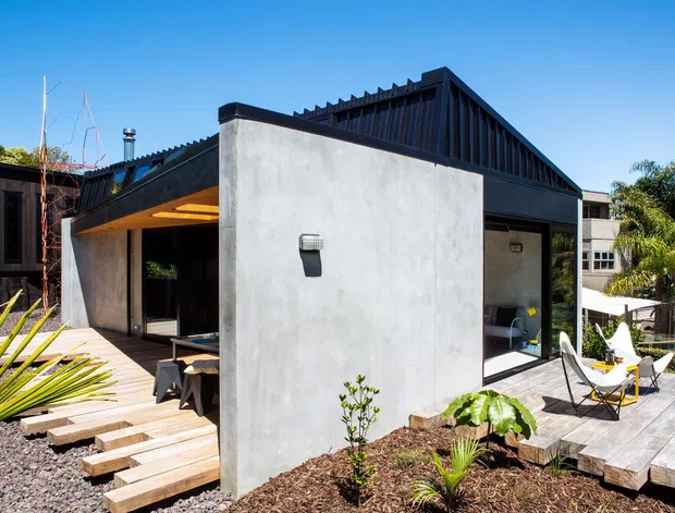 Exterior view of a contemporary single-storey residence with strong geometric volumes, a smooth grey concrete wall, dark timber-clad sections, and a steeply pitched ribbed roof form. Broad timber decking and stepped timber pavers connect to open glazed living areas, with landscaped planting around the house and outdoor seating on the deck.