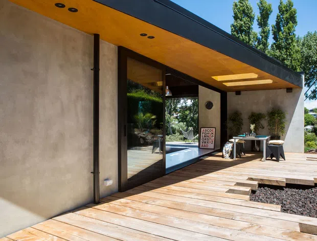 Exterior view of a contemporary single-storey building with smooth rendered walls, dark flat roof edges, warm timber soffit, and large dark-framed glazing opening onto a broad timber deck. The deck leads through landscaped gravel and planting toward an entrance or outdoor living area, with a small table and stools visible beneath the covered overhang.