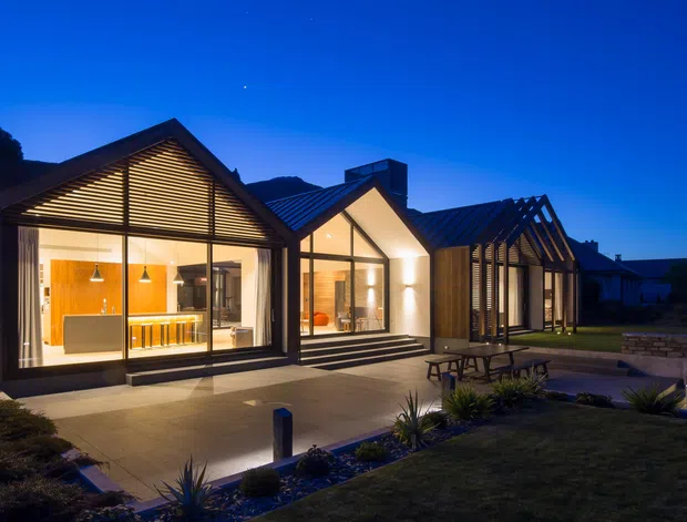 Dusk exterior view of a contemporary single-storey residence composed of linked gabled volumes. The home has dark metal pitched roofs, extensive floor-to-ceiling glazing, warm illuminated interiors, timber-toned wall finishes, and a broad paved terrace with outdoor seating and landscaped planting. A pool or water feature is visible at the right edge.