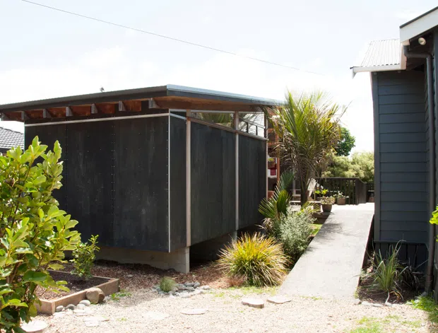 Exterior view of a compact contemporary outbuilding or extension clad in dark weathered panels, with a low-slung flat roof and clerestory-like openings beneath the eaves. A narrow concrete path runs between the structure and a dark blue-grey weatherboard house, surrounded by established subtropical planting and gravel landscaping.