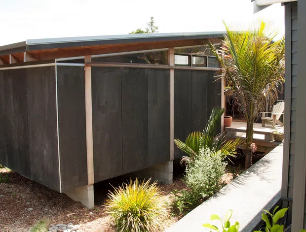 Exterior view of a compact dark-clad backyard structure or studio beside a contemporary home. The structure has a low-pitched metal roof with exposed timber soffit, dark vertical wall panels, pale timber framing, high clerestory-style glazing, and is raised slightly on concrete supports. A narrow paved path, timber steps, and established ornamental planting surround it.