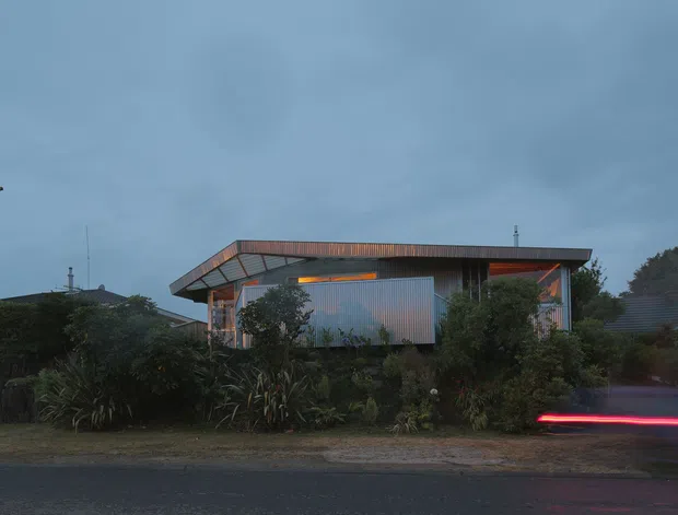 Exterior street view of a low-rise contemporary house at dusk. The building has a broad shallow roof with deep overhangs, a boxy composition, corrugated metal cladding, large glazed openings, and warm linear lighting beneath the eaves. Dense established planting screens much of the lower facade, while a blurred vehicle light trail passes along the road in the foreground.