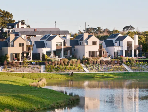 A row of contemporary multi-storey residential homes arranged along landscaped waterfront grounds. The buildings feature white rendered facades, dark pitched metal roofs, projecting balconies, large windows, and private planted courtyards. A pond occupies the foreground, with shared lawns, paths, ornamental grasses, and structured garden beds between the homes and water.