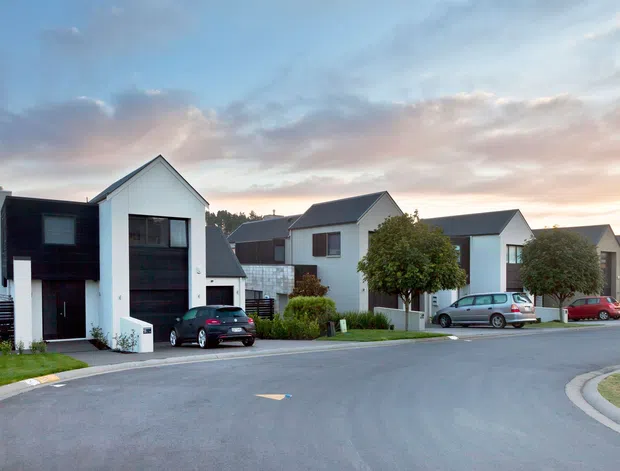 A suburban residential streetscape at dusk showing a row of contemporary detached homes with crisp gabled roof forms, white rendered walls, dark vertical cladding, and attached or integrated garages. Driveways, parked cars, small landscaped front gardens, and a curved paved road are visible.