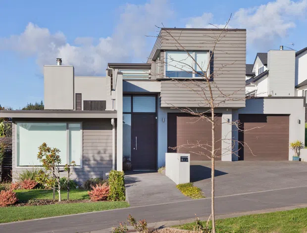 A modern multi-level residential home viewed from the street, featuring a crisp geometric facade, horizontal weatherboard-style cladding, large glazed openings, a projecting upper storey, a dark entry door, and an integrated double garage. The front garden includes lawns, young trees, and low ornamental planting, with neighbouring homes visible in the background.