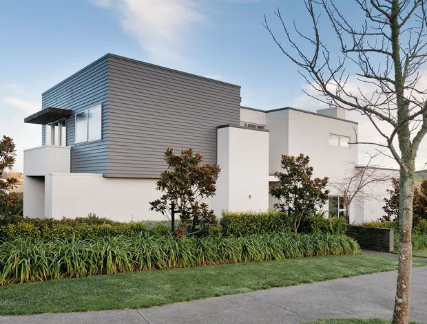 Exterior view of a contemporary multi-level residential house with a clean geometric composition. The building combines white rendered volumes with a prominent horizontal grey cladding box, large modern windows, and a projecting flat canopy. Low-maintenance landscaped planting, lawn, street verge and a mature deciduous tree frame the facade.