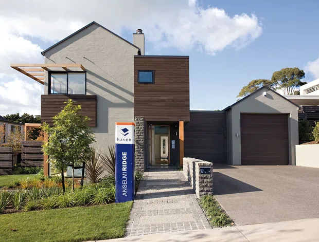 Exterior view of a contemporary multi-level residential home with a gabled rendered facade, dark horizontal timber cladding, projecting glazed upper-level balcony or sunroom, and an attached single-car garage. A paved pedestrian path leads through landscaped planting to the recessed entry, while a broad driveway occupies the right side.