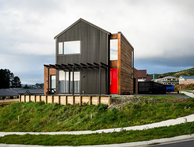 A contemporary multi-level house on a sloping suburban site, featuring a steep gable-roofed upper volume clad in dark vertical ribbed metal, a timber-clad side wall, brick lower-level elements, extensive glazing, and a striking red entrance panel. A pergola-covered glazed outdoor area projects from the lower level, with landscaped grass and a curved concrete walkway in the foreground.