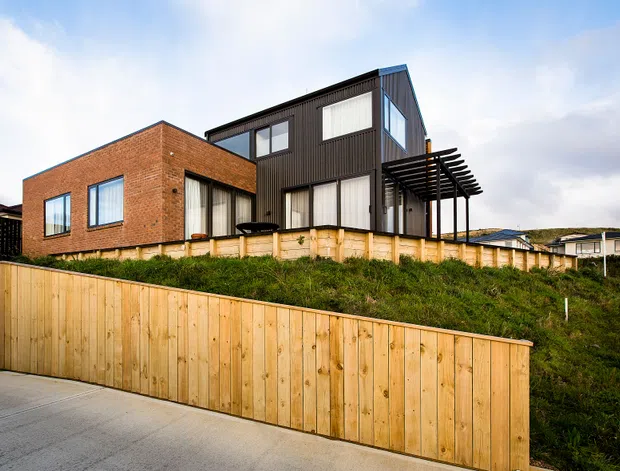 Exterior architectural photograph of a contemporary multi-level house on a sloping suburban site. The building combines a red brick volume with a dark vertically clad upper volume, large rectangular windows, and a timber pergola over a terrace. Timber retaining and boundary fencing line the foreground, with neighbouring homes visible in the background.