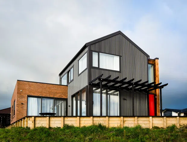 Exterior view of a contemporary multi-level residential house with a steep gabled roof, dark vertically ribbed cladding, extensive glazing, a projecting timber pergola, and a contrasting red entry door. A lower brick-clad wing and pale timber retaining wall sit in front of the house on a grassy site.