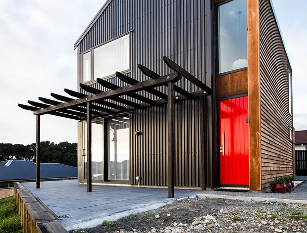 Exterior view of a compact modern multi-level house on a raised, landscaped site. The building has dark vertically ribbed metal cladding, warm timber cladding and framing, large windows, a bright red entry door, and a black timber pergola over a paved terrace. The ground is rocky and lightly landscaped, with neighbouring rooftops and trees in the background.