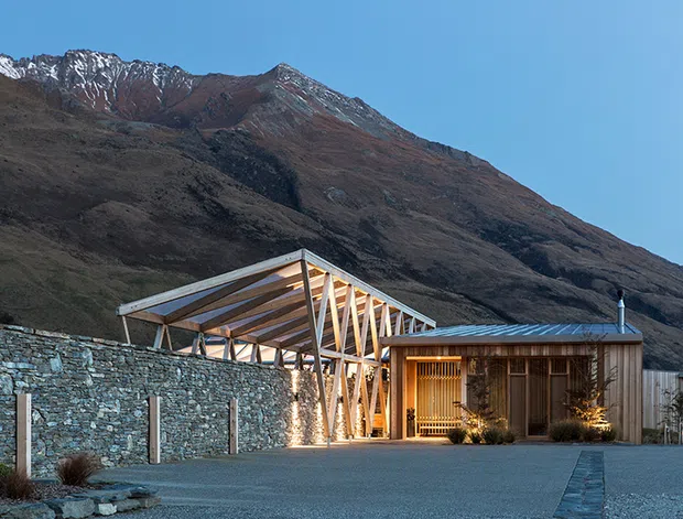 A low-rise mountain residence or lodge set against a steep, rugged alpine mountain backdrop. The building combines a transparent timber-framed pavilion with exposed triangular roof structure and a more enclosed timber-clad wing with a shallow metal roof. A dry-stone retaining wall, gravel forecourt, planted borders and warm interior lighting define the exterior setting.