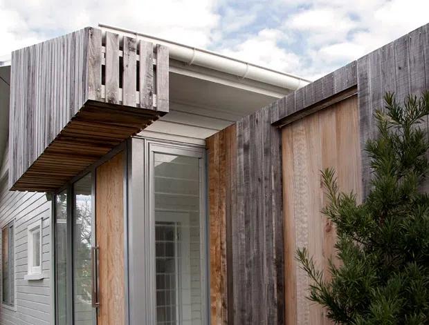 Exterior view of a compact contemporary building entrance, featuring weathered vertical timber screening, light grey horizontal cladding, a glazed door, and a projecting timber-clad canopy. Dense landscaping partially obscures the lower facade, while a bright cloudy sky is visible above.