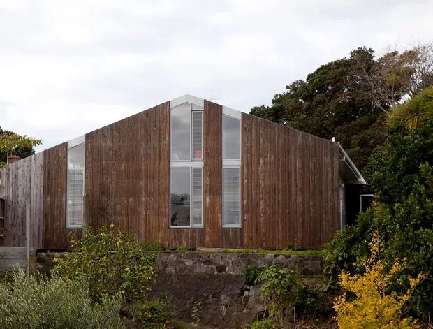 Exterior view of a compact contemporary timber-clad house set on a raised dark stone retaining wall. The facade has vertical weathered wood boards, narrow vertical glazing panels with louvred sections, and a simple asymmetrical pitched roof. Dense vegetation surrounds the building.