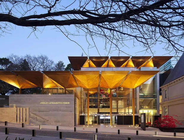 Exterior view of a contemporary cultural or commercial building at dusk, featuring a dramatic illuminated folded-metal canopy with repeated angular roof forms over a tall glazed entrance. A masonry-clad wing and ornate historic-looking adjacent building frame the composition, with bollards and a paved forecourt in front.