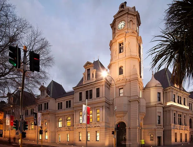 Street-level dusk view of an ornate historic civic or commercial building on a city corner. The cream-coloured masonry facade features classical detailing, tall sash windows, projecting wings, steep slate roofs, dormers, turrets and a prominent illuminated clock tower. Traffic lights, street signage and mature trees frame the urban streetscape.