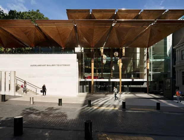 Exterior view of the Auckland Art Gallery Toi o Tāmaki, showing a prominent glass entrance facade, pale stone-clad gallery volume, and a dramatic canopy formed by repeated timber roof planes and supporting columns. The image is taken from a paved urban forecourt with pedestrian bollards and surrounding mature trees.