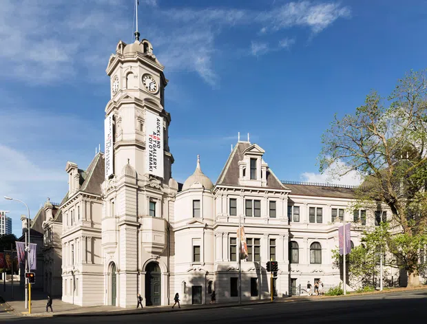 Street-level exterior photograph of a grand historic civic or institutional building in an urban setting. The pale stone or rendered façade features ornate classical detailing, a prominent clock tower with a pointed roof and flagpole, steep gables, pilasters, arched openings, and decorative cornices. Banners hang from the tower, with pedestrians and a broad street in the foreground.