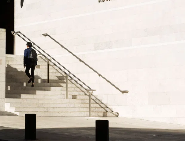 Exterior view of the Auckland Art Gallery entrance, featuring a broad pale stone-clad wall with raised lettering, a wide flight of steps, stainless-steel handrails, dark paving, bollards, and a glazed upper terrace beneath a projecting roof. A person is ascending the stairs.