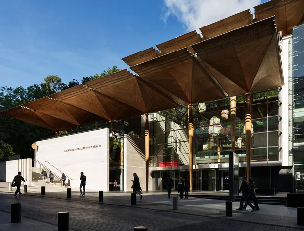 Exterior view of a contemporary cultural or commercial building with a large timber canopy composed of repeated angular folded roof sections, supported by slender timber columns. A glazed multi-storey entrance facade and a pale rendered wall frame the paved public forecourt, with pedestrians and bollards visible.
