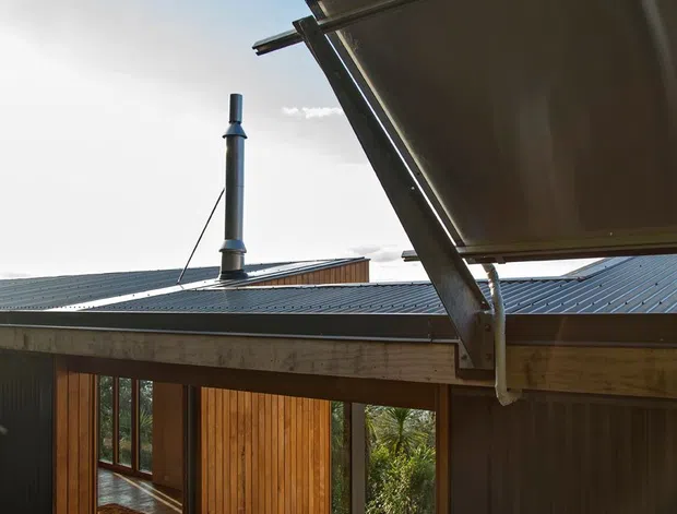 Exterior architectural view of a compact timber-and-metal building with a corrugated metal pitched roof, exposed roof structure, and a large raised awning or canopy in the foreground. Vertical timber cladding, dark metal wall panels, concrete blockwork, and extensive glazed openings surround a timber deck and planted courtyard-like setting. A roof flue is visible against a bright blue sky.