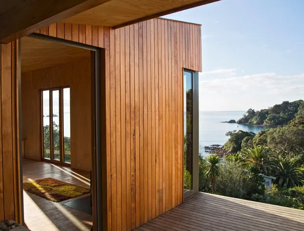 Exterior architectural view of a contemporary timber-clad coastal dwelling, showing a covered outdoor deck, vertical wood cladding, large glazed openings, and warm timber-lined interior spaces opening toward a sheltered bay and dense coastal vegetation.