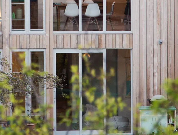 Exterior view of a two-storey residential building clad in vertical pale timber boards, with extensive aluminium-framed glazing and sliding doors. Through the upper glazing, a dining area with a table, modern chairs, pendant lights and kitchen joinery is visible. The lower level opens onto a small paved outdoor area, partially obscured by foreground planting. A corrugated metal roof edge is visible at the top.