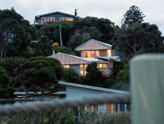Exterior view of a multi-level residential property set within dense coastal vegetation. Several interconnected volumes feature steep pitched roofs, timber or weatherboard-style cladding, extensive glazing, and warm interior lighting. The foreground is partially obscured by a blurred balustrade or fence, while a taller dark-clad upper dwelling is visible behind the lower structures.