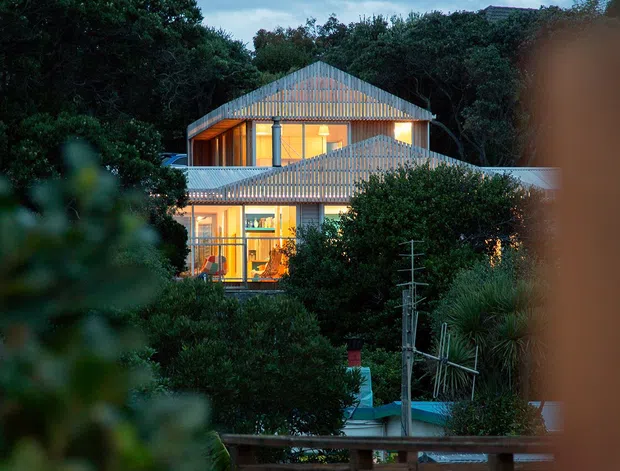 Exterior view of a multi-level contemporary residence framed by dense greenery, photographed at dusk. The house features large illuminated glazed openings, layered corrugated metal gable roofs, and timber-toned exterior elements. A pool or water feature, fencing, and utility infrastructure are visible in the foreground, with blurred vegetation framing the image.
