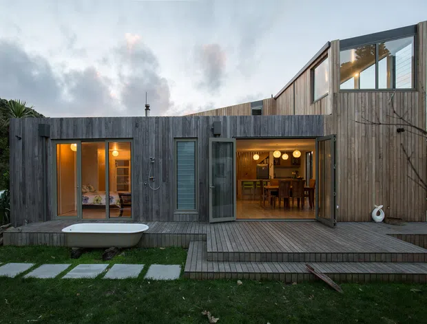 Exterior view of a compact contemporary timber-clad house with a low, angular roofline and large glazed openings. A raised timber deck fronts the home, with a freestanding outdoor bathtub, lawn stepping stones, and warm interior lighting visible through the windows and open doors. The upper-level glazing and varied volumes create a modern, sculptural composition.