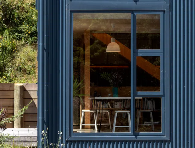 Close-up exterior view of a contemporary dark blue corrugated-metal-clad building with a tall multi-pane window. The glazing reflects surrounding vegetation and sky while revealing a warmly lit interior with a dining table, stools, bookshelves, plants, and a pendant light. A grey concrete-block base, timber retaining structure, and planted garden surround the facade.
