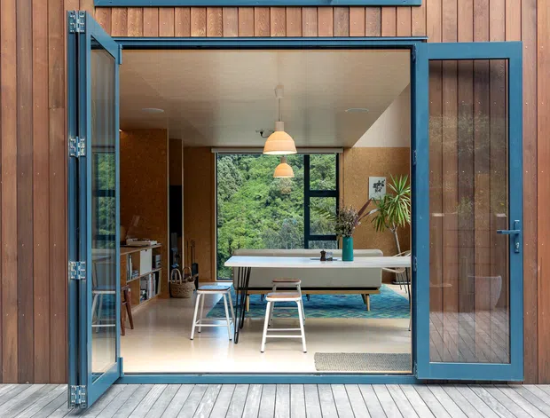 View from a timber-clad exterior through wide-open blue-framed glazed doors into a bright, carefully furnished living and dining space. The room features warm timber wall finishes, a pale floor, pendant lights, a pool table or large central table, indoor plants, and a large window overlooking dense greenery. A pale timber deck runs across the foreground.