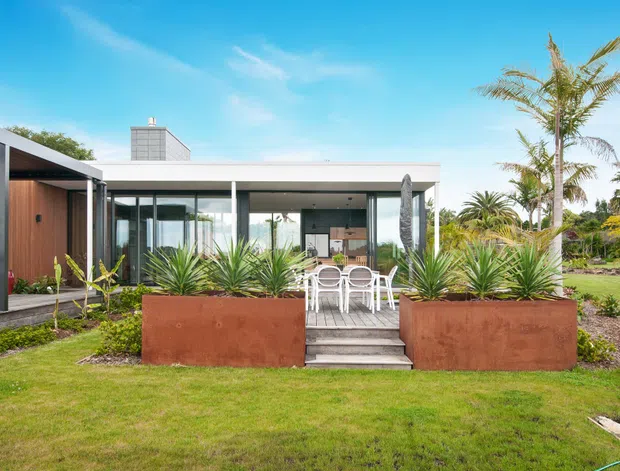 Exterior view of a low, single-storey contemporary home with extensive floor-to-ceiling glazing, a broad flat roof, dark slender frames, and warm timber wall cladding. A paved patio with white outdoor dining furniture sits between large rusted steel planter boxes, surrounded by tropical planting and lawn.