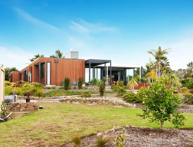 A single-storey contemporary residential building set within a landscaped garden. The home combines warm vertical timber cladding with extensive dark-framed glazing and a simple flat-roofed form, including a projecting covered terrace or verandah. Stone retaining walls, gravel garden beds, lawns, and established subtropical planting surround the house.