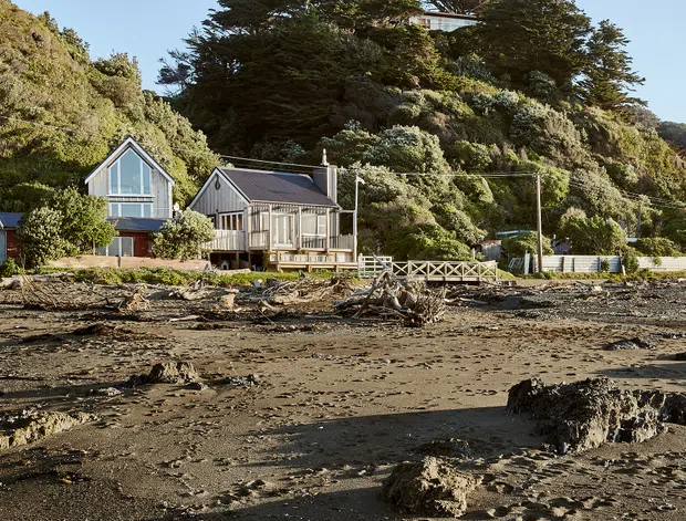 Exterior view of a group of compact coastal houses positioned directly behind a broad rocky and sandy foreshore. The buildings feature steep gable roofs, vertical weatherboard-style cladding, large glazed gable ends, and timber-framed windows and decks. Dense coastal vegetation and rugged hills rise behind the homes, while exposed mudflats and driftwood dominate the foreground.