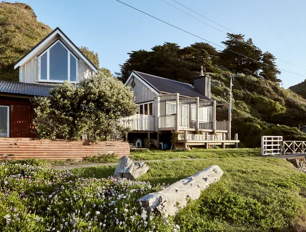 Exterior view of two small timber-clad coastal cottages on a grassy, rocky site. The buildings feature steep gabled roofs, large gable-end glazing, raised decks, corrugated metal roofing, and simple verandah structures. Dense coastal vegetation and exposed rock surround the homes, with a timber pedestrian bridge or deck railing at right and overhead utility lines visible.