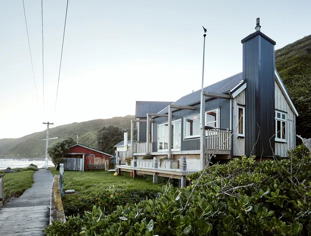 Exterior view of a narrow, multi-gabled coastal house clad in dark vertical metal siding, with a tall chimney, covered porch, timber deck and large windows. The home sits on a rugged sloping coastal site beside a simple outbuilding, with a path leading toward the shoreline. Overhead utility lines and low vegetation reinforce the remote coastal setting.