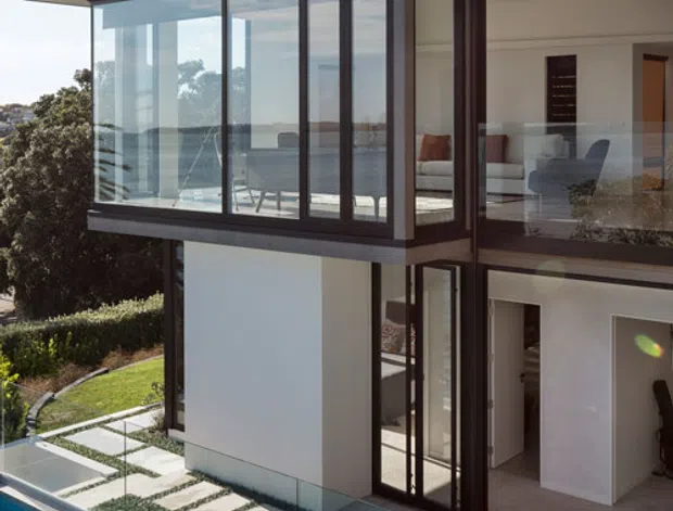 Exterior view of a contemporary multi-storey residence with strong horizontal roof planes, dark framed floor-to-ceiling glazing, and white rendered walls. A swimming pool with frameless glass fencing and a patterned paved walkway occupies the foreground, while furnished living areas are visible through the glazing. The home overlooks a broad waterfront or coastal vista.