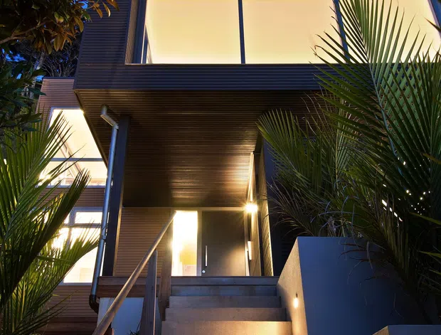 Dusk exterior view looking up a concrete stairway toward the entrance of a modern multi-level residence. The facade combines dark horizontal cladding with extensive floor-to-ceiling glazing, a deeply recessed entry and soffit lighting. Tropical palm planting frames the stairs, while warm interior and pathway lighting contrasts with the blue evening sky.