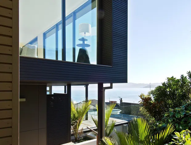 Exterior architectural photograph of a contemporary elevated coastal home. The view focuses on a projecting upper-level room with large floor-to-ceiling glazing, dark horizontal cladding, timber decking, and an open lower level supported by posts. Tropical planting surrounds the house, with a pool and neighbouring rooftops visible below and a broad water view beyond.