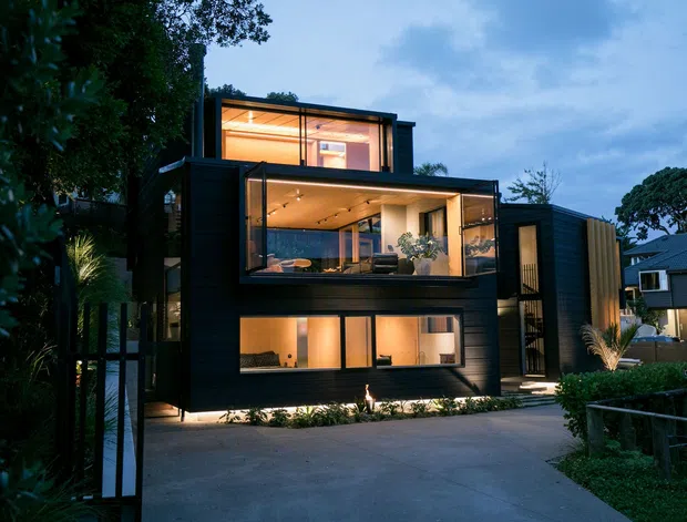 A contemporary multi-level dark-clad residence photographed at dusk, with extensive floor-to-ceiling glazing revealing warmly lit living spaces. The home has a strong cubic composition, projecting upper-level volumes, a flat roofline, landscaped planting, and a broad concrete driveway at the front.