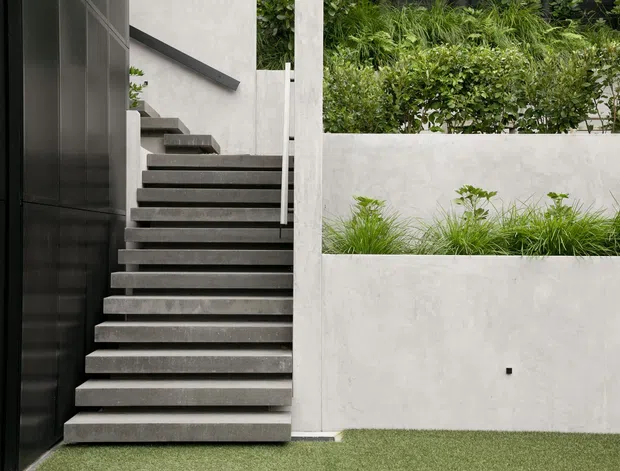 A minimalist exterior entrance featuring a dark metal-clad wall, a straight flight of broad concrete stairs with a slim metal handrail, and rendered concrete garden retaining walls with integrated planting. The composition is clean and contemporary, with a small lawn in the foreground and dense greenery behind the courtyard walls.