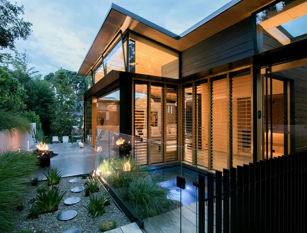 Contemporary two-storey house viewed from a landscaped outdoor approach at dusk. The building features strong angular roof planes, extensive floor-to-ceiling glazing, dark horizontal cladding, warm timber soffits, and timber louvres. A gravel garden with grasses, stepping stones, illuminated planting, water or reflective surfaces, and an outdoor fire feature frames the entrance.