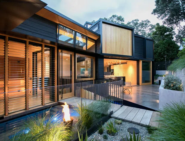 Contemporary multi-level home exterior viewed from a landscaped courtyard, featuring dark cladding, extensive glazing, warm timber screening, projecting box forms, and an open entertaining area. A paved terrace, railing, planted garden beds, and a small illuminated water or fire feature occupy the foreground.