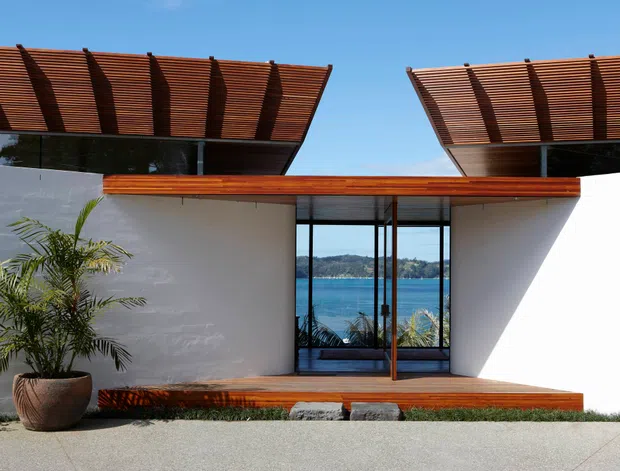A contemporary waterfront residential facade with crisp white rendered walls, expansive central glazing, and warm timber roof canopies projecting on both sides. The deep slatted timber screens create strong horizontal and linear shadows, while a timber deck and low built-in bench frame the entrance. Blue water and a wooded shoreline are visible through the glazing, with a potted palm in the foreground.