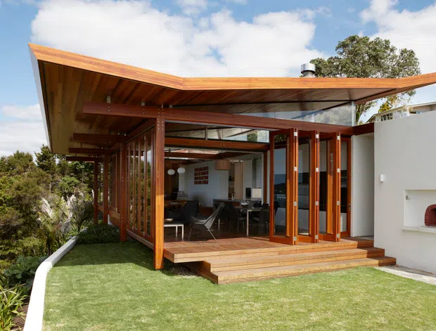 Exterior view of a contemporary single-storey home with a dramatic low-pitched projecting timber roof and extensive floor-to-ceiling glazing. A timber-framed, glass-enclosed living and dining area opens onto a raised timber deck with broad steps. The white rendered wall includes a built-in outdoor pizza oven or fireplace, while landscaped lawn and planting surround the structure.